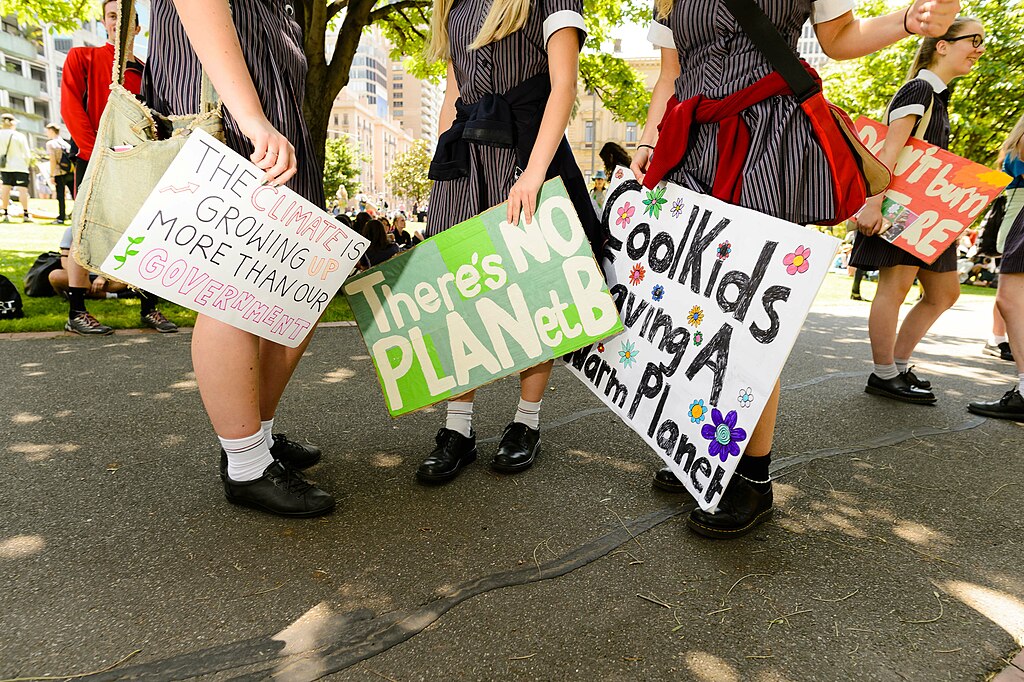 Students in school uniforms holding posters at the School Strike for Climate in 2018. The signs read "The climate is growing more than our government", "There's NO PLANet B" and "Cool kids saving a warm planet". 